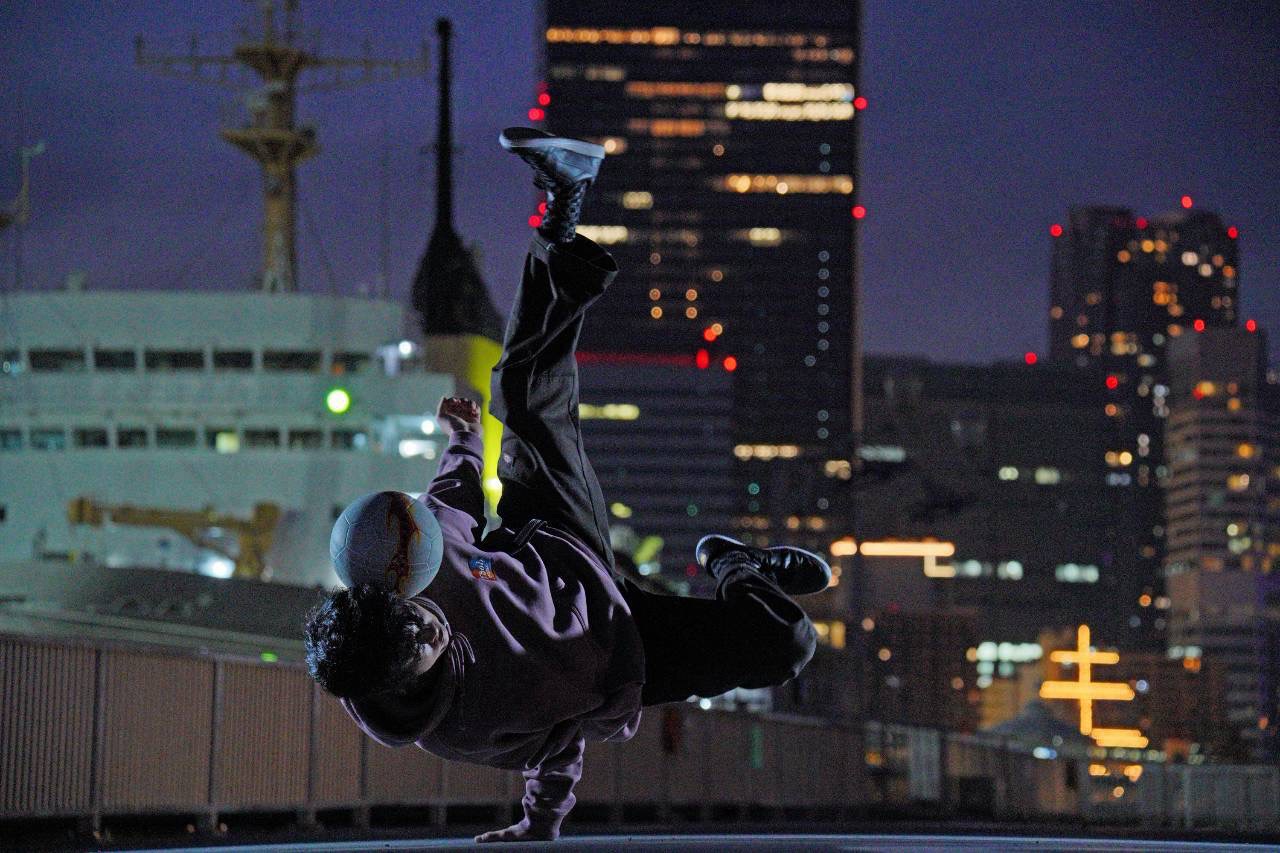 夜景を背に、片手で体を支えて逆立ちの体勢をとるSITZ