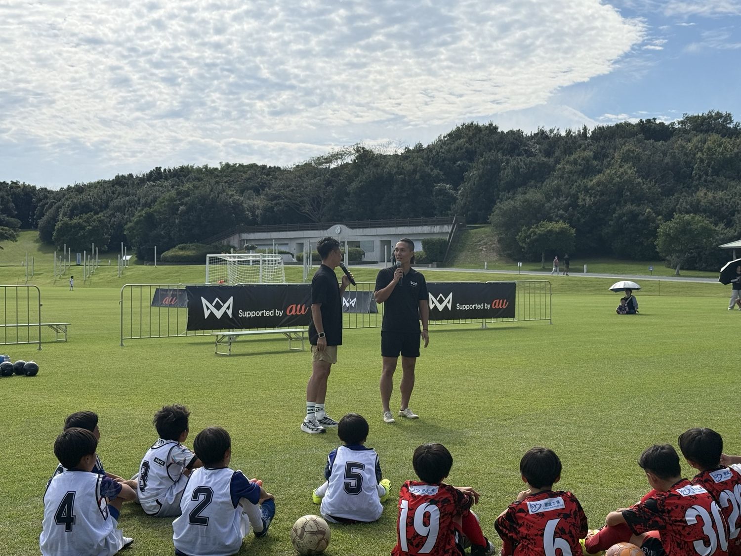 芝生に座る子どもたちの前で、久保裕也さんとマイクで話すMCおのしん