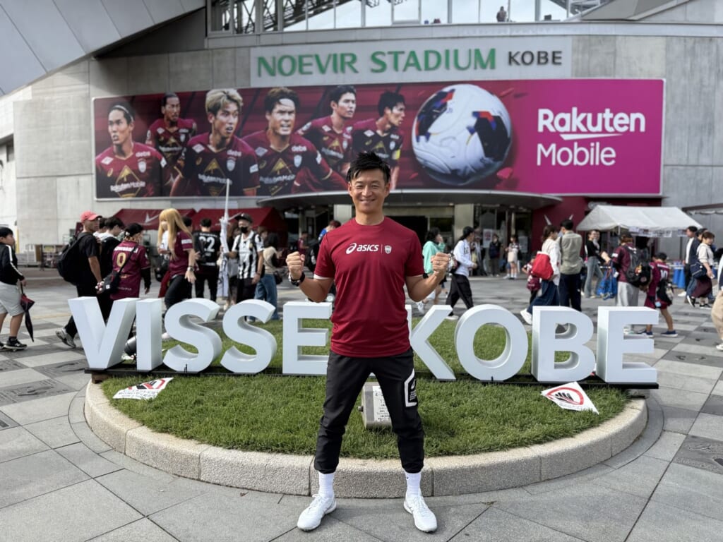 ノエビアスタジアム神戸の前、VISSEL KOBEの文字の前で笑顔でガッツポーズをするMCおのしん