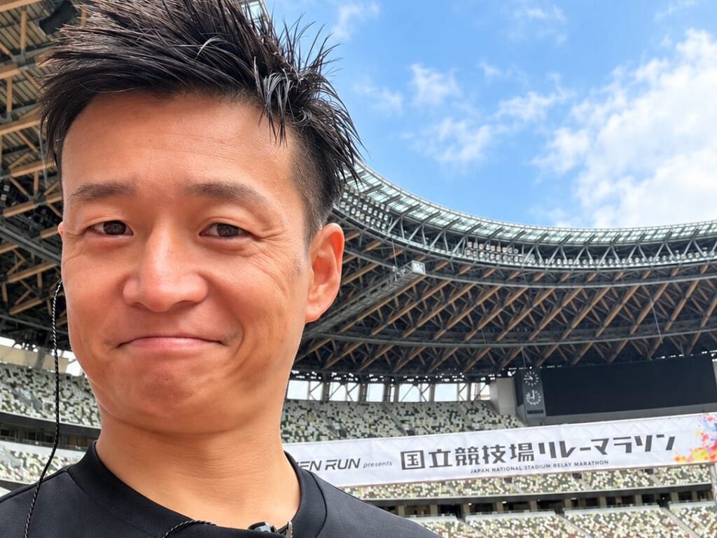 国立競技場のトラック脇で、スタンドと大会バナーを背にしたMCおのしん